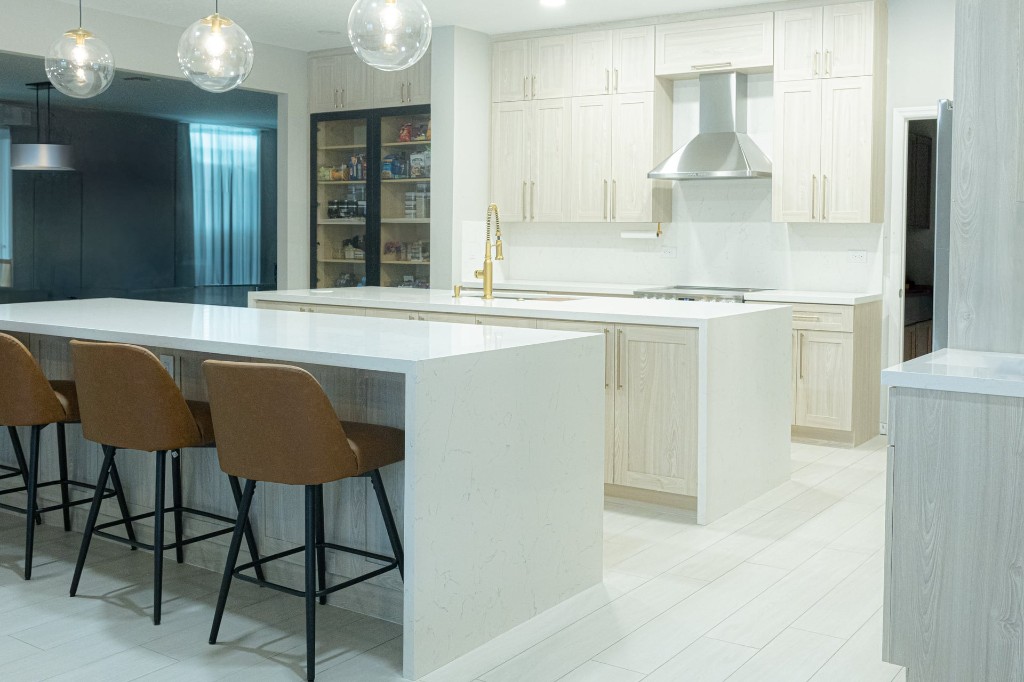 Modern custom kitchen with dual white waterfall islands, light wood cabinetry, and glass-door pantry
